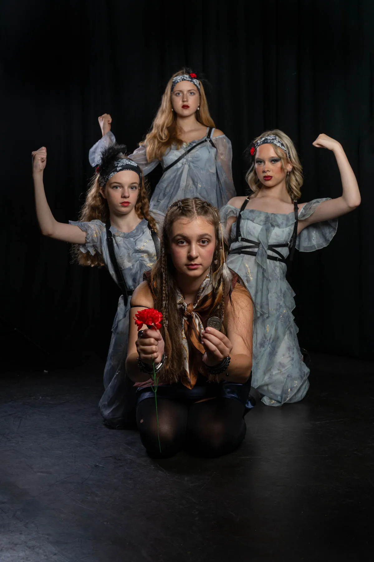 Four cast members in Hadestown costume, three in pale floral dresses with fists raised behind one kneeling with a red carnation and a coin