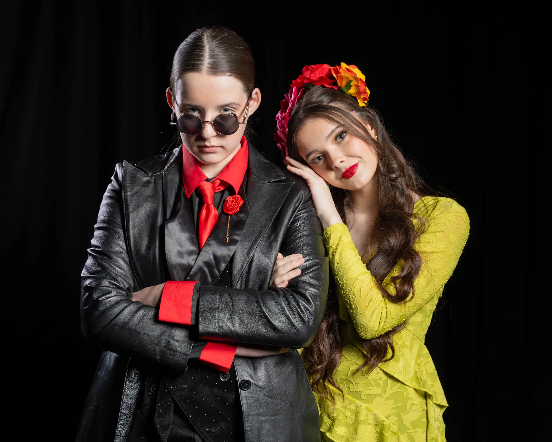 Two cast members in Hadestown costume, one in a black leather coat, red tie and round sunglasses, the other in a yellow-green dress and flower crown