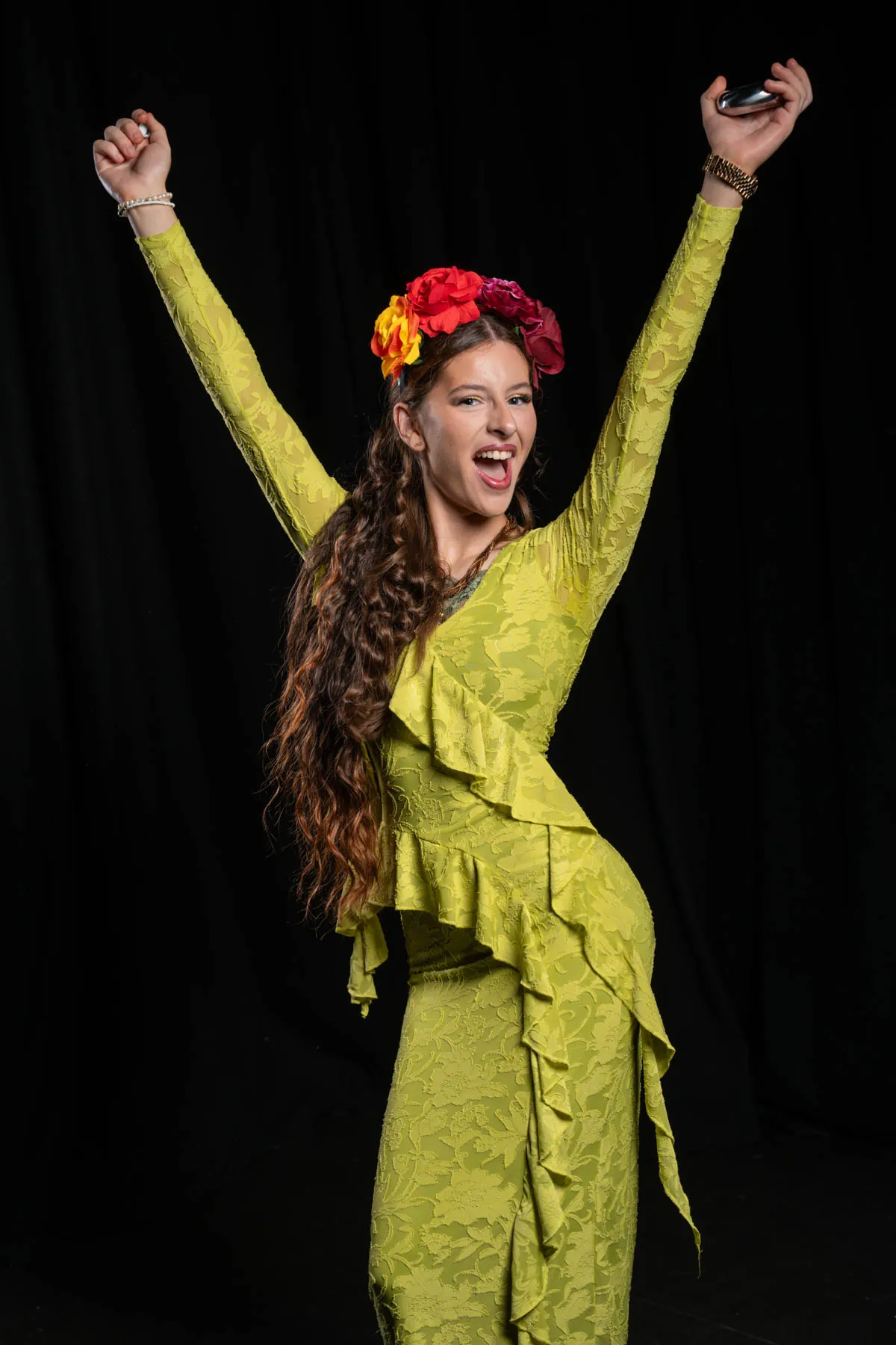 A cast member in a yellow-green ruffled dress and flower crown with both arms raised