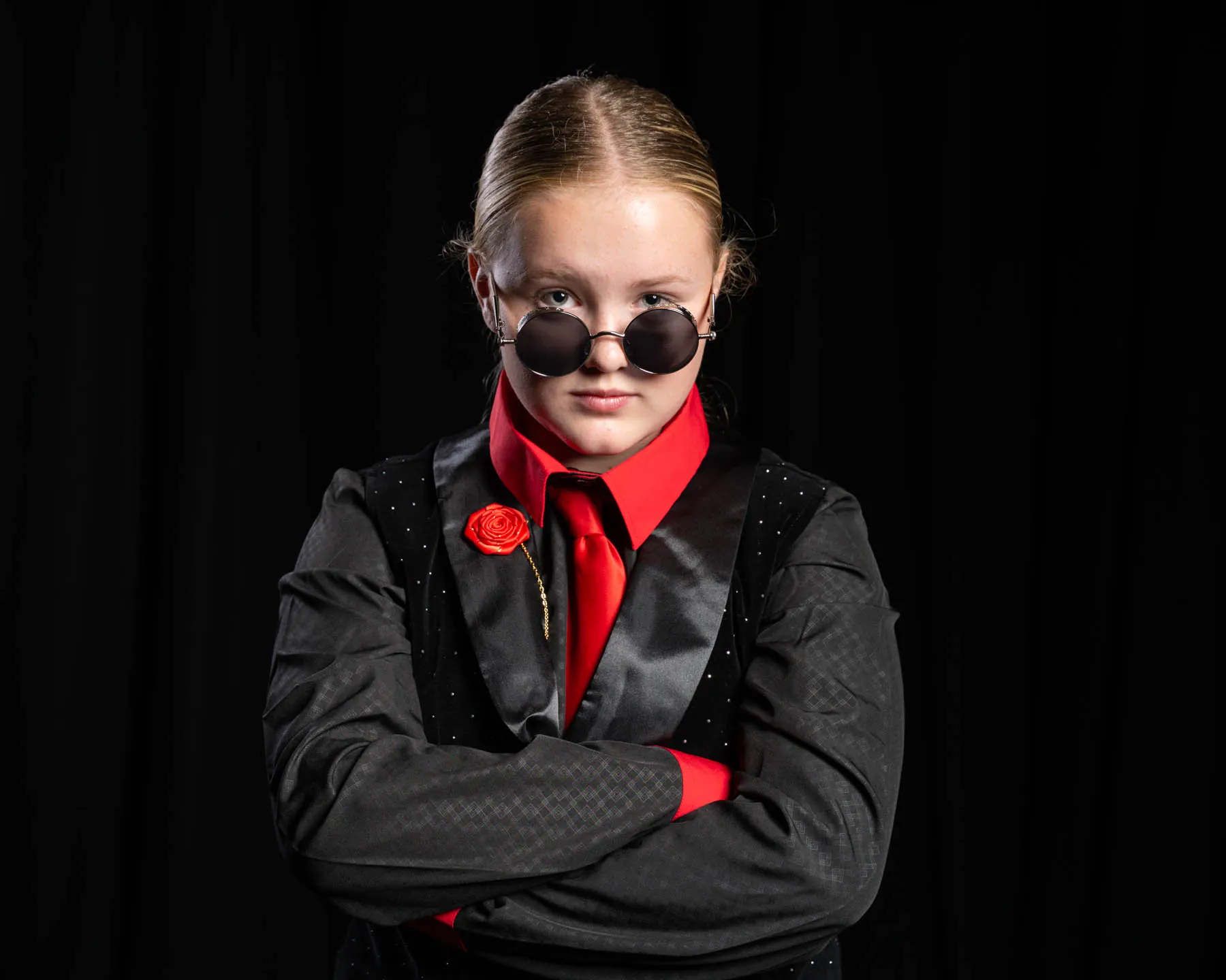 A cast member in a black suit, red shirt and tie and round sunglasses with arms folded