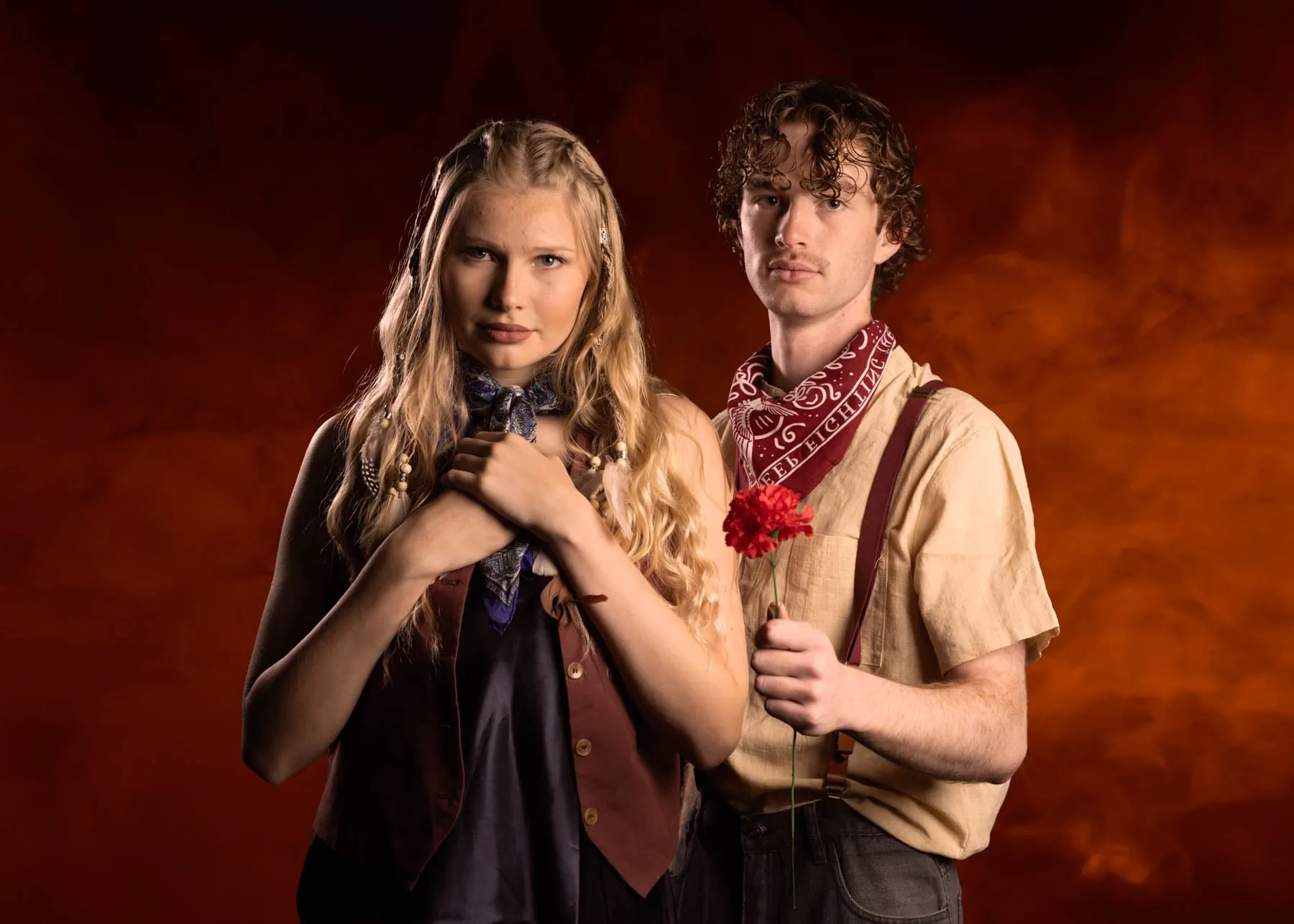 Two cast members against a smoky orange backdrop, one with braided hair and a neck scarf, the other holding a red carnation