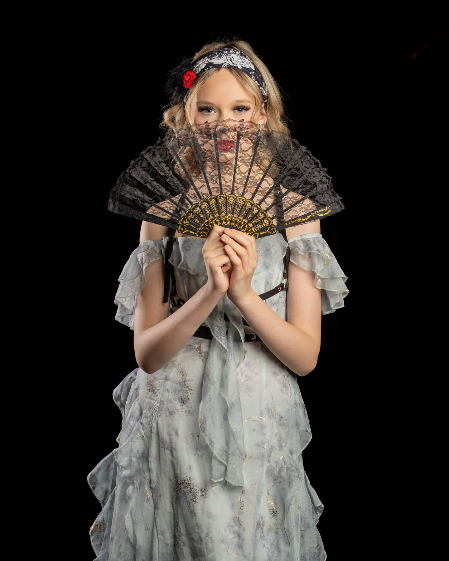 A cast member in a pale ruffled dress holding a black lace fan in front of their face